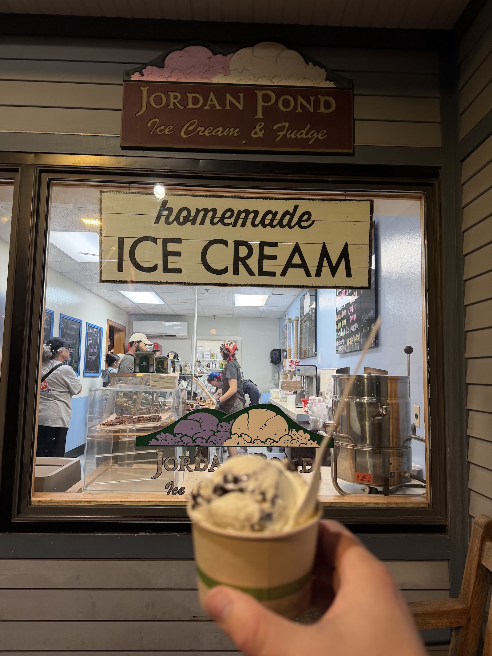 The carved “Jordan Pond Ice Cream & Fudge” sign above a “homemade ice cream” window sign in the evening, a cup of cookies-and-cream in the foreground.
